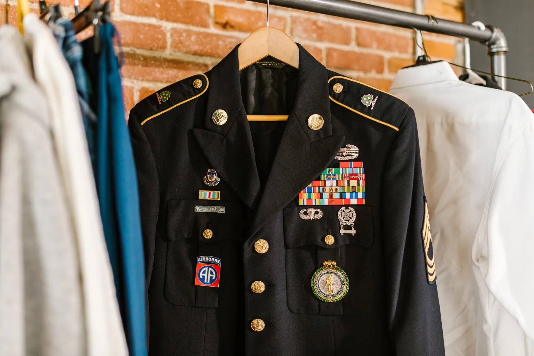 A dark navy dress uniform jacket on a wooden hanger, chest ribbons, badges and chevrons visible, brick wall behind.