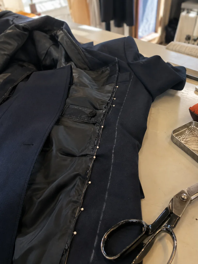 A navy suit jacket inside out on a work table, the side seam opened and re-pinned to taper the waist, a chalk line along the new seam.