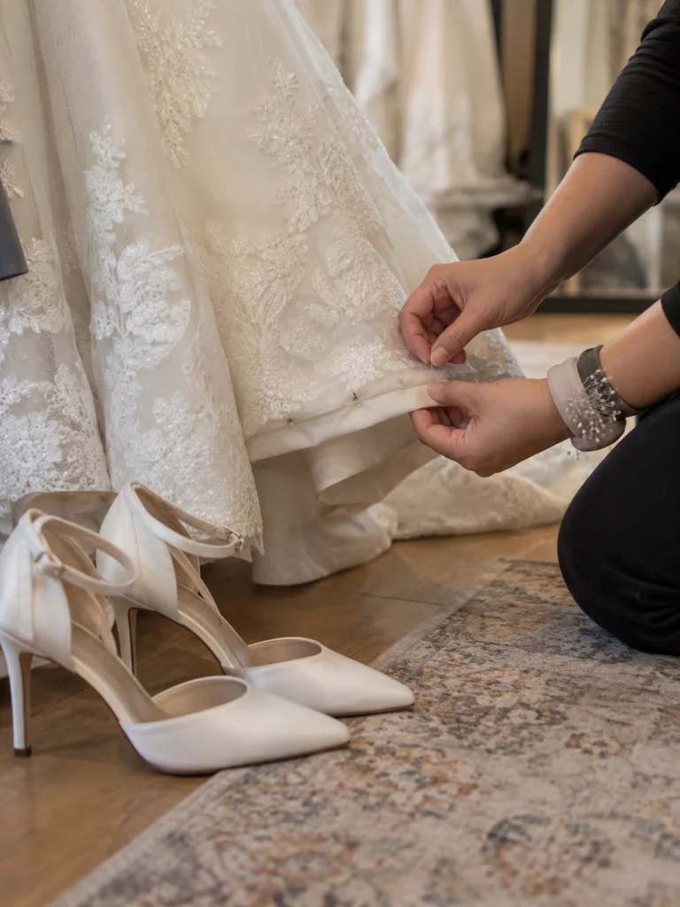 Ivory heels on a wooden floor beside the hem of a wedding gown, the hem pinned to length just above the shoe.