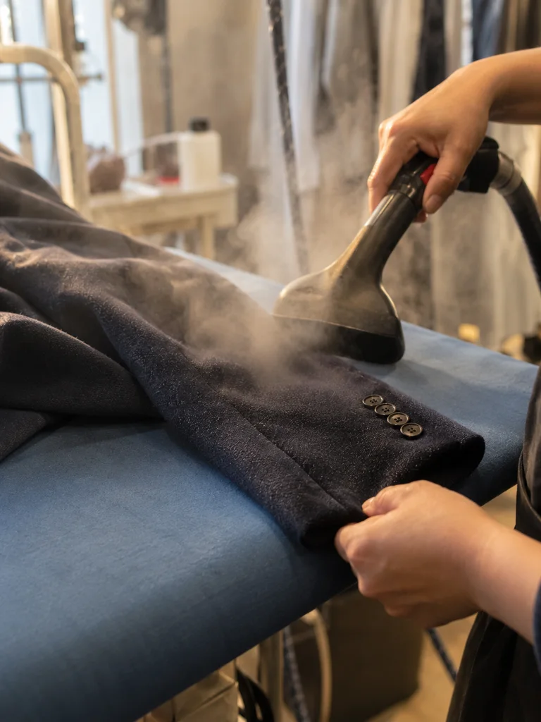 A navy wool blazer sleeve on a pressing table under a hand-held steamer, steam lifting the nap.