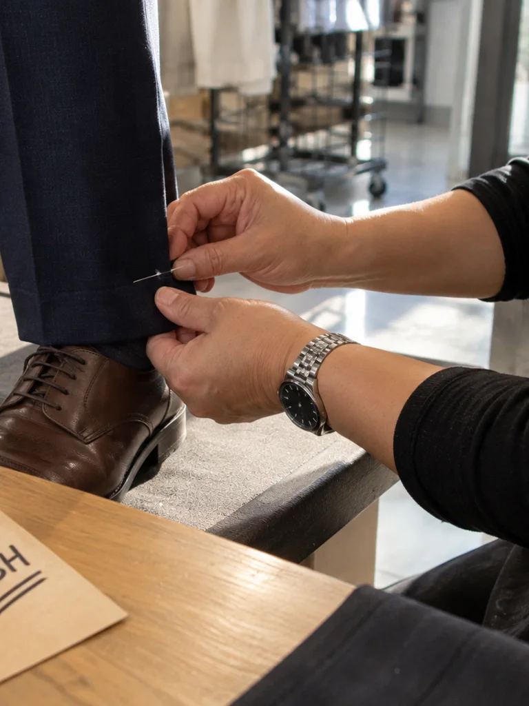 Hands pinning a trouser hem at a customer’s ankle, a wristwatch on the seamstress, a kraft rush tag on the table.